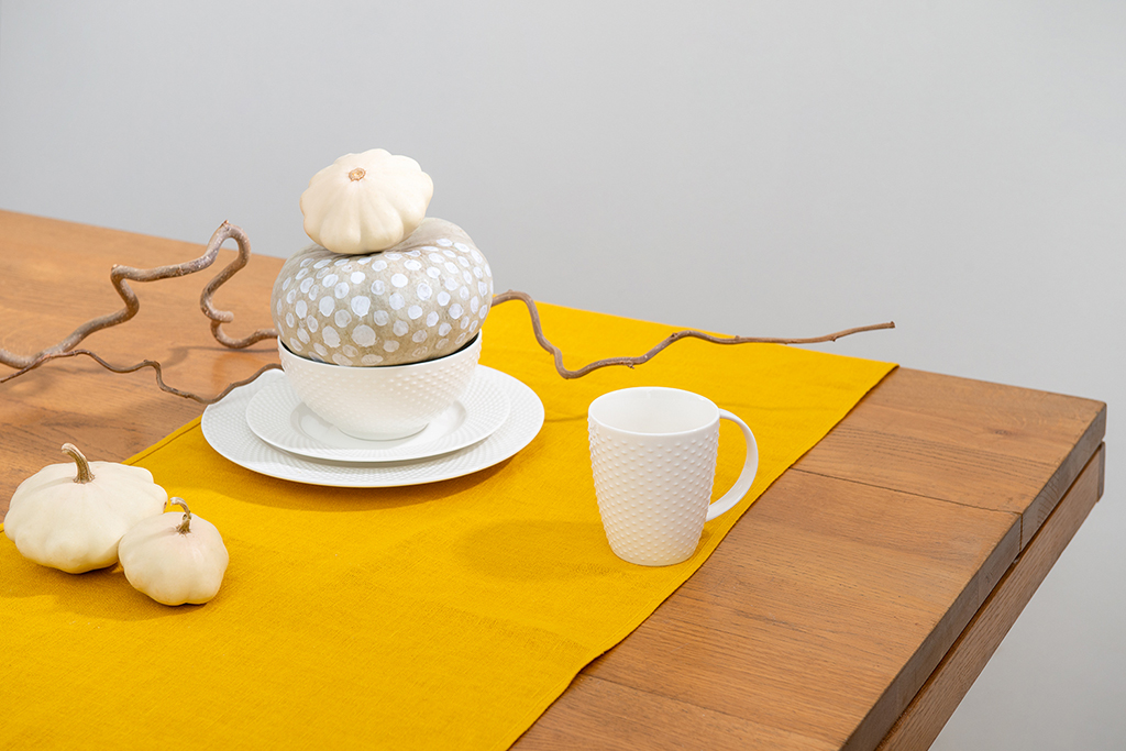На фото товары из коллекции Essential бренда Tkano: дорожка на стол из стираного льна горчичного цвета и кружка белого цвета с фактурным рисунком